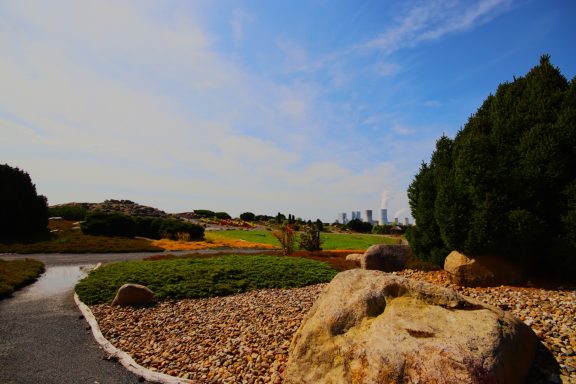 Landschaft mit Grünflächen, Bäumen und Felsen unter einem blauen Himmel.