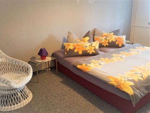 Schlafzimmer mit Doppelbett, bunten Kissen und einem Stuhl neben einem Tisch.